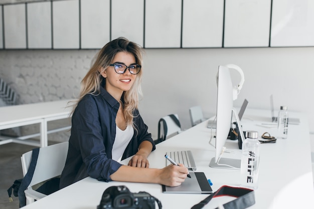 Foto gratuita attraente designer femminile utilizzando tablet per lavoro, seduto in ufficio con interni luminosi