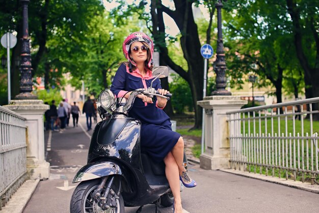 Привлекательная женщина в платье сидит на мотоскутере в осеннем парке.