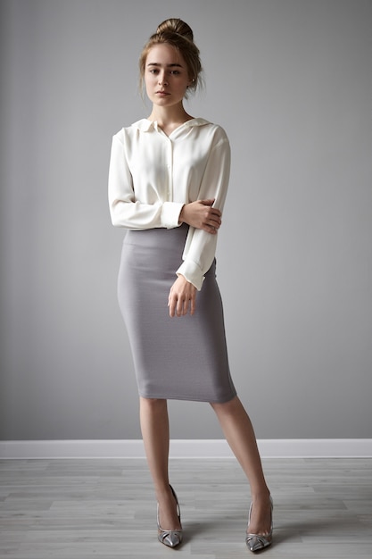 Attractive female employee with hair knot posing isolated against gray wall background, having serious look, dressed in elegant formal clothes. Vertical shot of thoughtful young businesswoman