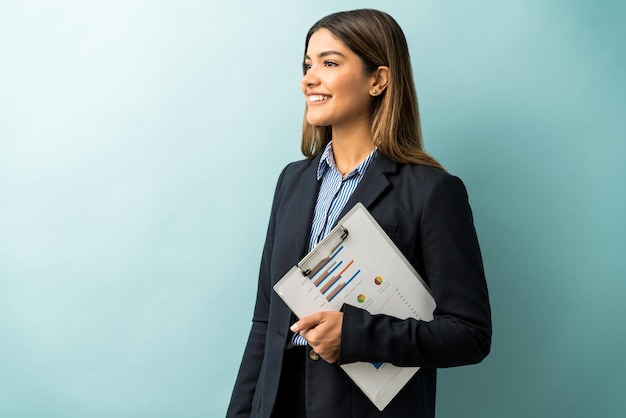 Attractive female entrepreneur holding clipboard with graph while looking away against colored background