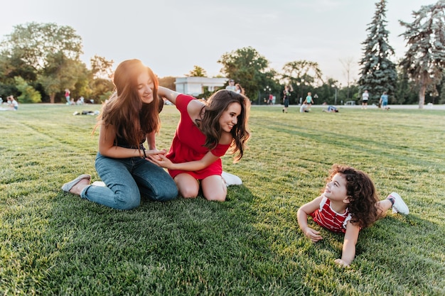 Attractive female friends posing on the grass with little kid. Pretty curly girl spending time with sisters in park.