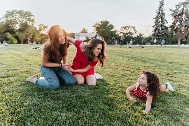 Attractive female friends posing on the grass with little kid. Pretty curly girl spending time with sisters in park.