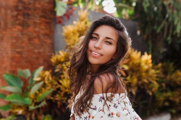 Attractive female green-eyed brunette in white top with bare shoulders looking at front with smile on street with tropical plants