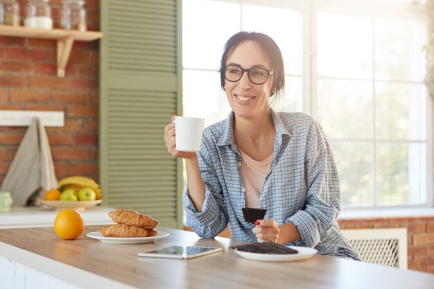 Attractive female has happy expression enjoys morning coffee with sweet delicious croissants and chocolate