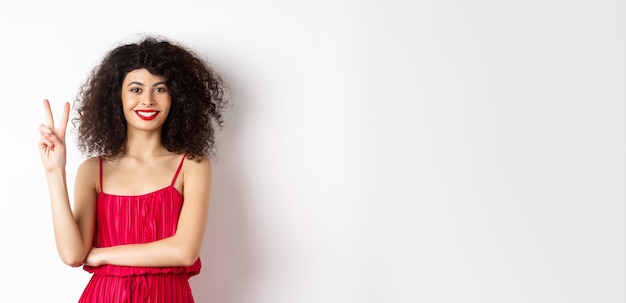 Attractive female model in red dress and makeup showing number two and smiling standing over white b