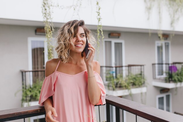 Привлекательная женская модель с короткими волнистыми волосами зовет друга из отеля. прекрасная молодая женщина в розовой блузке разговаривает по телефону, стоя на балконе.