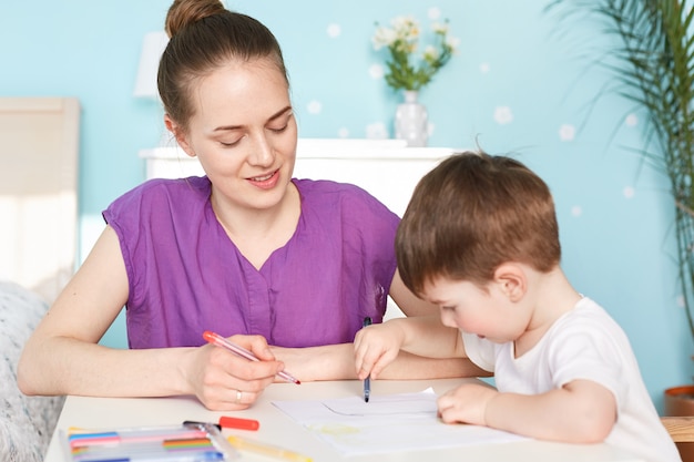 Attractive female mother sits near her little son who draws picture on blank piece of paper