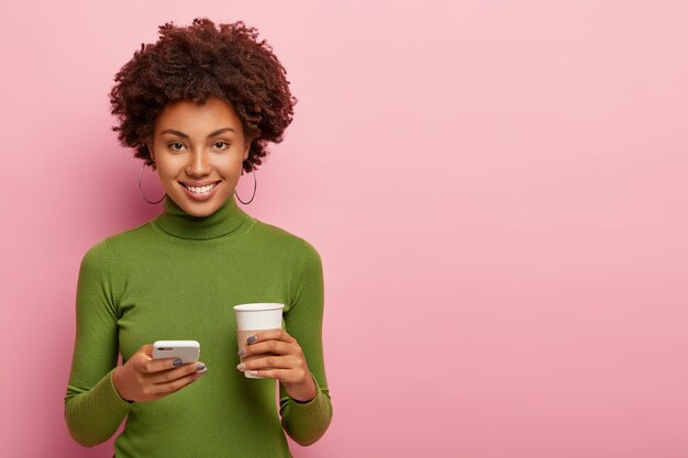 Привлекательная женщина с довольным выражением лица, держит мобильный телефон и кофе с собой, одетая в зеленую одежду, отправляет текстовое сообщение, общается в чате онлайн, изолирована на розовой стене