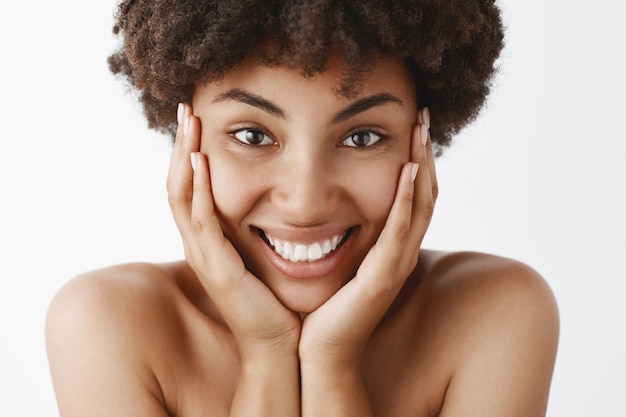 Привлекательная женственная и натуральная афроамериканская молодая женщина с вьющимися волосами и чистой чистой кожей, трогающая лицо и широко улыбаясь от волнения и счастья, позирует обнаженной над серой стеной