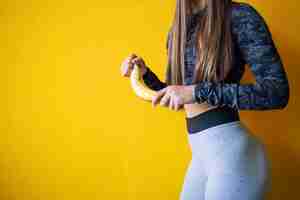 Free photo attractive fitness women in sportswear peeling off banana fruit against yellow wall