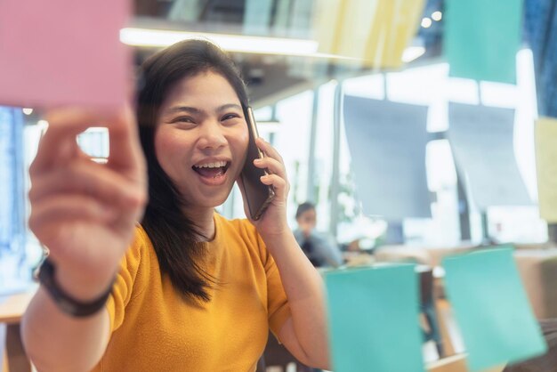 매력적인 프리랜서 아시아 여성 창의적인 사람은 사려 깊고 신선함을 갖춘 코워킹 스페이스의 새로운 라이프스타일에서 스마트폰 손 포인트 종이 메모 알림과 함께 캐주얼한 노란색 천으로 대화를 나눕니다.