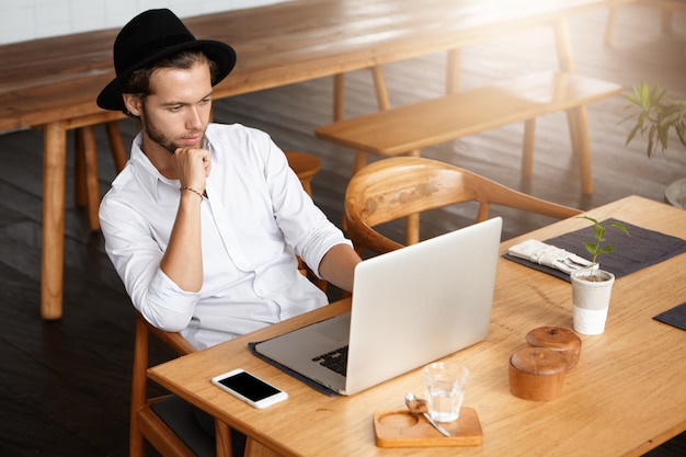 Attractive freelancer dressed in white shirt working remotely sitting at wooden table in front of open laptop computer and looking at screen with thoughtful confident expression, leaning on his elbow