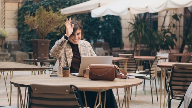 Attractive friendly businesswoman greeting while talking with colleagues by video call in cafe outdoor Young female entrepreneur using laptop