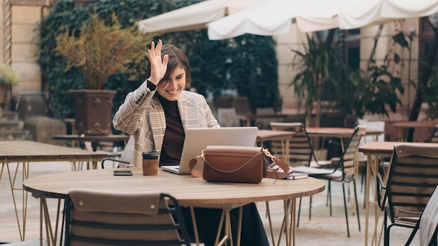 ノートパソコンを使用してカフェ屋外の若い女性起業家でビデオ通話で同僚と話している間魅力的なフレンドリーな実業家の挨拶