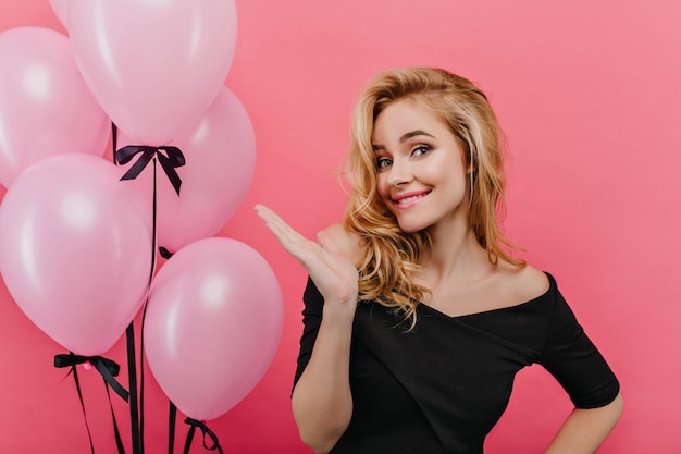 Attraente ragazza divertente con capelli biondi che gode del servizio fotografico del partito. interessato modello femminile in abito nero in piedi vicino a palloncini e sorridente.