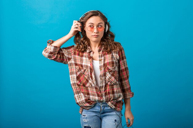 Attractive funny woman with surprised suspicious face expression looking aside listening to music in headphones in checkered shirt and jeans isolated on blue studio background