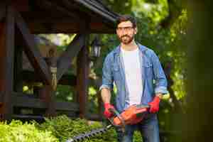 Free photo attractive gardener posing while trimming conifer bush by electric hedge clippers in sunny day