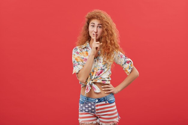 Attractive ginger female asking to be quiet while standing against red background