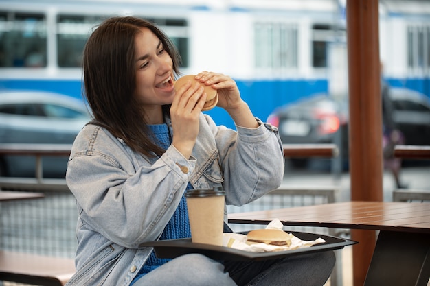 캐주얼 스타일의 매력적인 소녀는 여름 테라스에 앉아 커피와 함께 햄버거를 먹는다