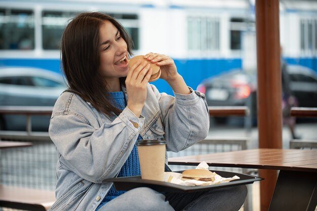 カジュアルなスタイルの魅力的な女の子は、夏のテラスに座ってコーヒーとハンバーガーを食べる