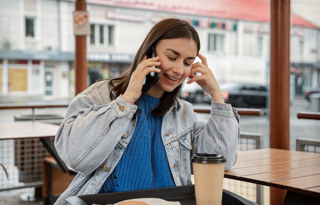 カジュアルなスタイルの魅力的な女の子は、カフェのテラスに座って、電話で話します