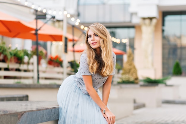 Attractive girl girl with long blonde hair in blue tulle skirt leaning on concrete bench on street. She is looking to camera.