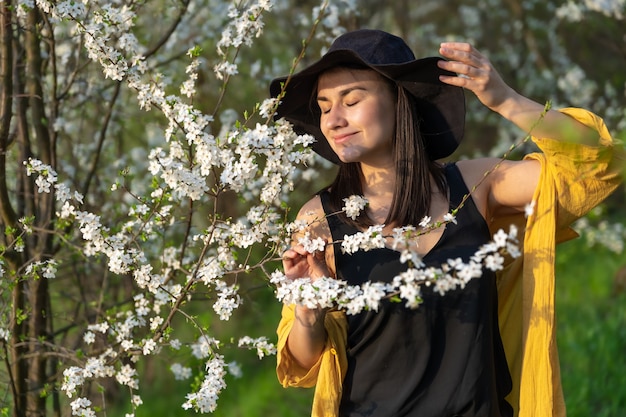 An attractive girl in a hat among blooming trees enjoys the smell of spring flowers