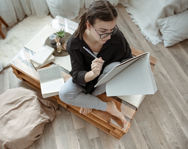 Attractive girl at home sitting with a notebook and pen among books top view.