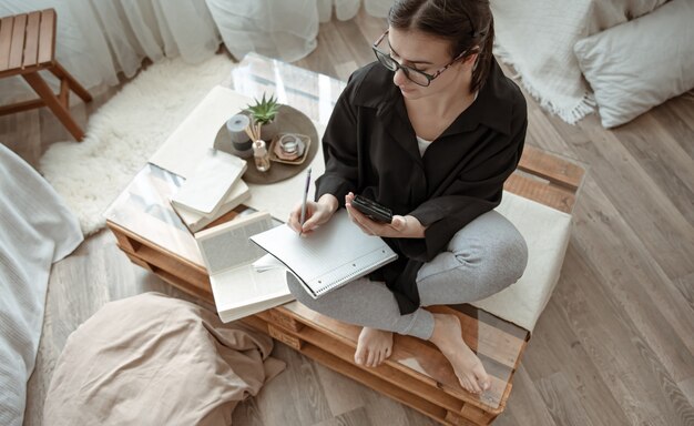 An attractive girl at home writes something in a notebook with a phone in her hands.