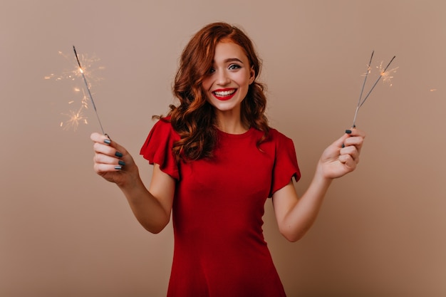 Attractive girl in red dress holding sparklers at new year party. Elegant ginger woman posing with bengal lights in christmas.