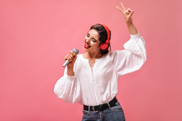 Attractive girl in red headphones singing in microphone on pink background.  Pretty lady with dark hair in white stylish blouse is listening to music.