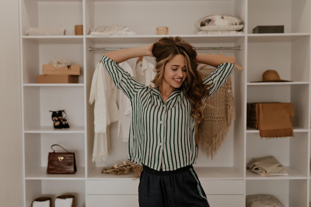 Attractive girl in silk pajamas ruffles hair in dressing room Cheerful woman in striped shirt smil