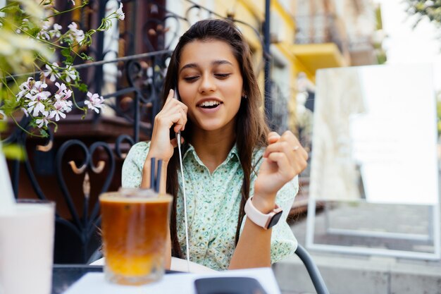Привлекательная девушка сидит на террасе уличного кафе на открытом воздухе, разговаривает со смартфоном, звонит другу