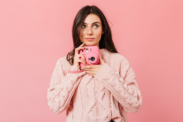 Attractive girl takes picture on instax camera. Lady in pink sweater looks up on isolated background.