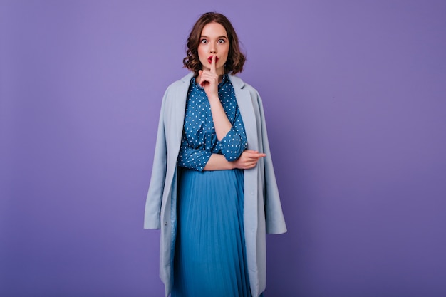 Attractive girl in vintage dress posing  with purple wall. Indoor photo of inspired curly lady in blue coat wants silence.