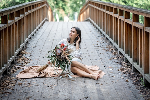 An attractive girl in a white dress sits on the bridge with a bouquet of exotic flowers.