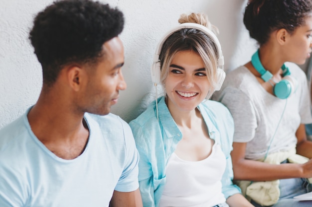 Attractive girl in white earphones looking with smile at her african friend in blue shirt. Black young man listening what his charming university mate talking.