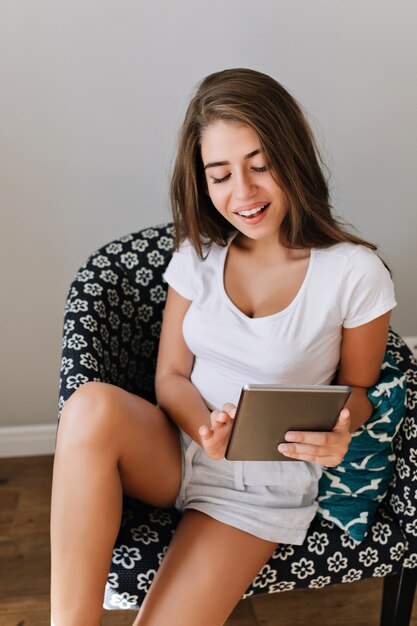 Attractive girl in white shirt  and shorts in chair. She smiling to tab in hands.