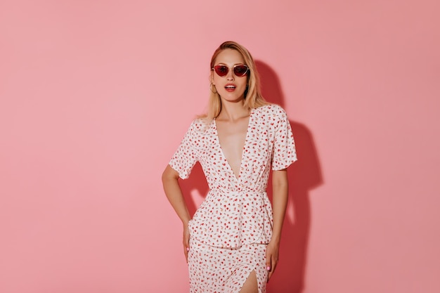 Attractive girl with blonde hair in red sunglasses and earrings in stylish white dress looking into camera on isolated pink backdrop