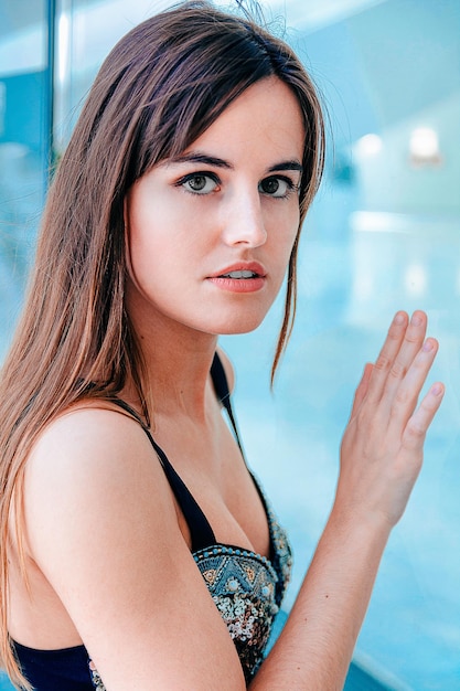 Attractive girl with brown long hair posing near the window with her hand on the glass