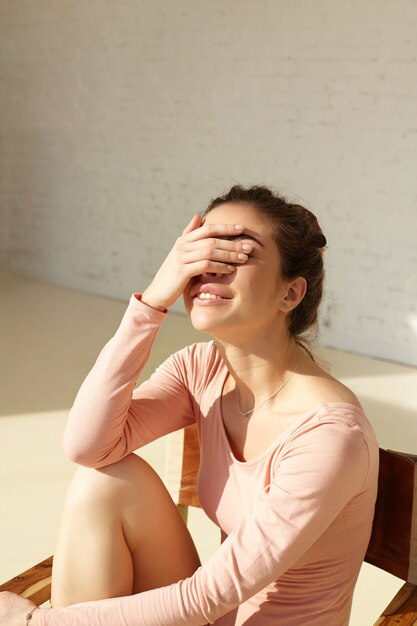 Attractive girl with cute smile covers face by hand squinting eyes in bright sun, having fun posing in modern interior at home. Smiling young fashion model enjoying the rest at home, copy space wall