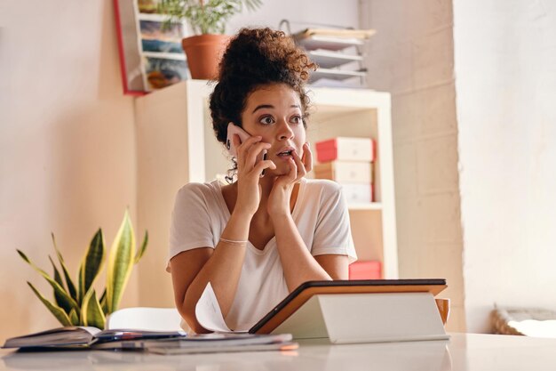 Attractive girl with dark curly hair sitting at the table with notebook and tablet amazedly looking aside talking on cellphone studying at cozy home