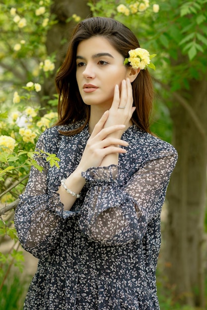 Attractive girl with flower in hair standing in blooming park looking forward touching face