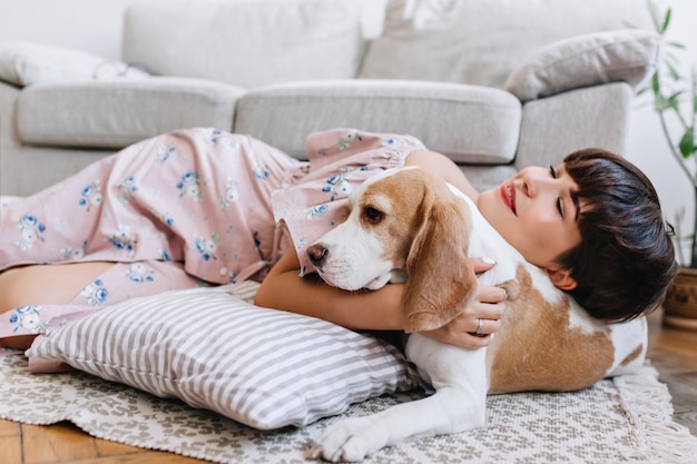 Attractive girl with happy face expression lies on carpet near beagle dog with light-brown ears