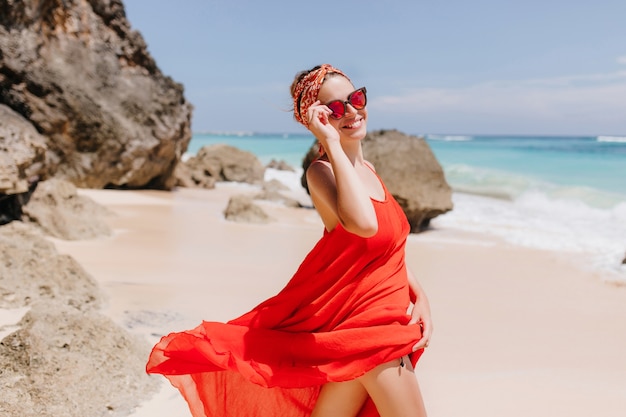 海の海岸を歩いて幸せな笑顔で魅力的な女の子。晴れた日のビーチでリラックスした赤いドレスとサングラスの洗練された女性モデル。