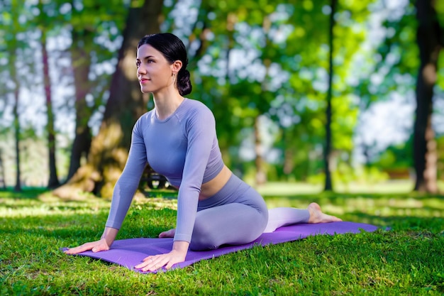 Attractive girl with long black hair dressed in purple doing yoga pose The Pigeon in the green park
