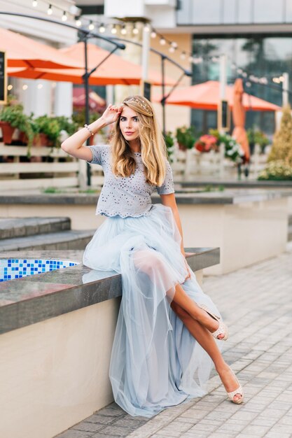 Attractive girl with long blonde hair in blue long tulle skirt sitting on terrace background. She keeps hand on head and looking to camera.
