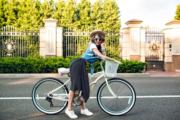Привлекательная девушка с длинными вьющимися волосами в шляпе позирует с велосипедом на дороге. Она носит длинную юбку, куртку, синие солнцезащитные очки. Она улыбается в камеру.