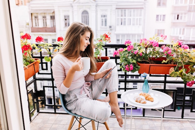 Attractive girl with long hair in pajama having breakfast on balcony in the morning in city. She holds a cup, reading on tablet.