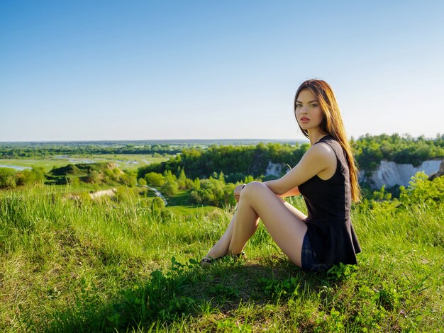 Attractive girl with long hair relaxes outdoor. Young woman sitting by a cliff outdoors on nature.  Female Model posing in a field on a sunny summer day.